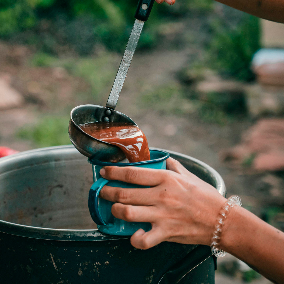 Person pouring a liquid from a ladle into a blue mug outdoors.