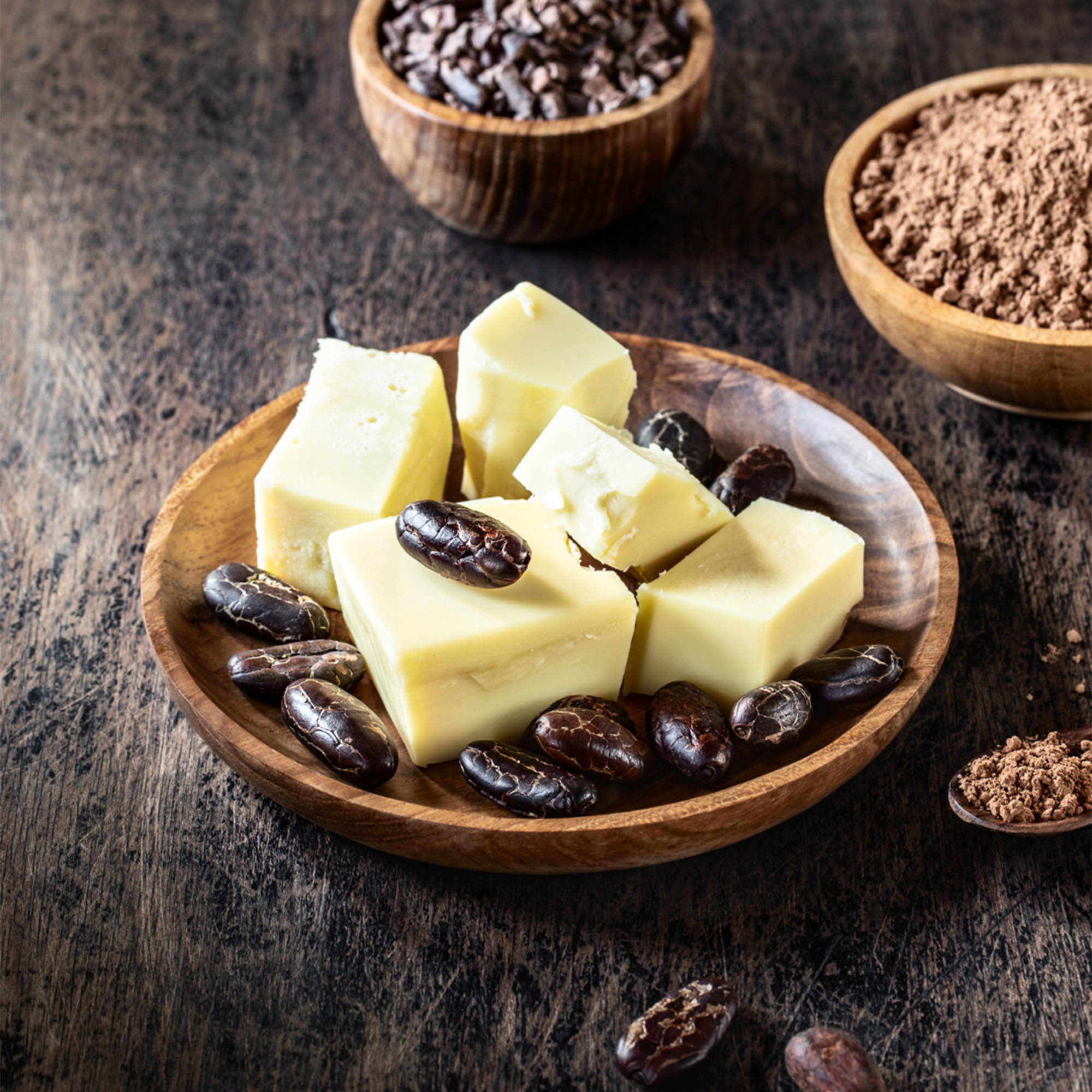 Wooden bowl with cubes of cacao butter, cacao beans, and cacao powder on a wooden surface