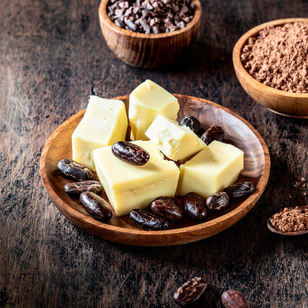Wooden bowl with cubes of cacao butter, cacao beans, and cacao powder on a wooden surface