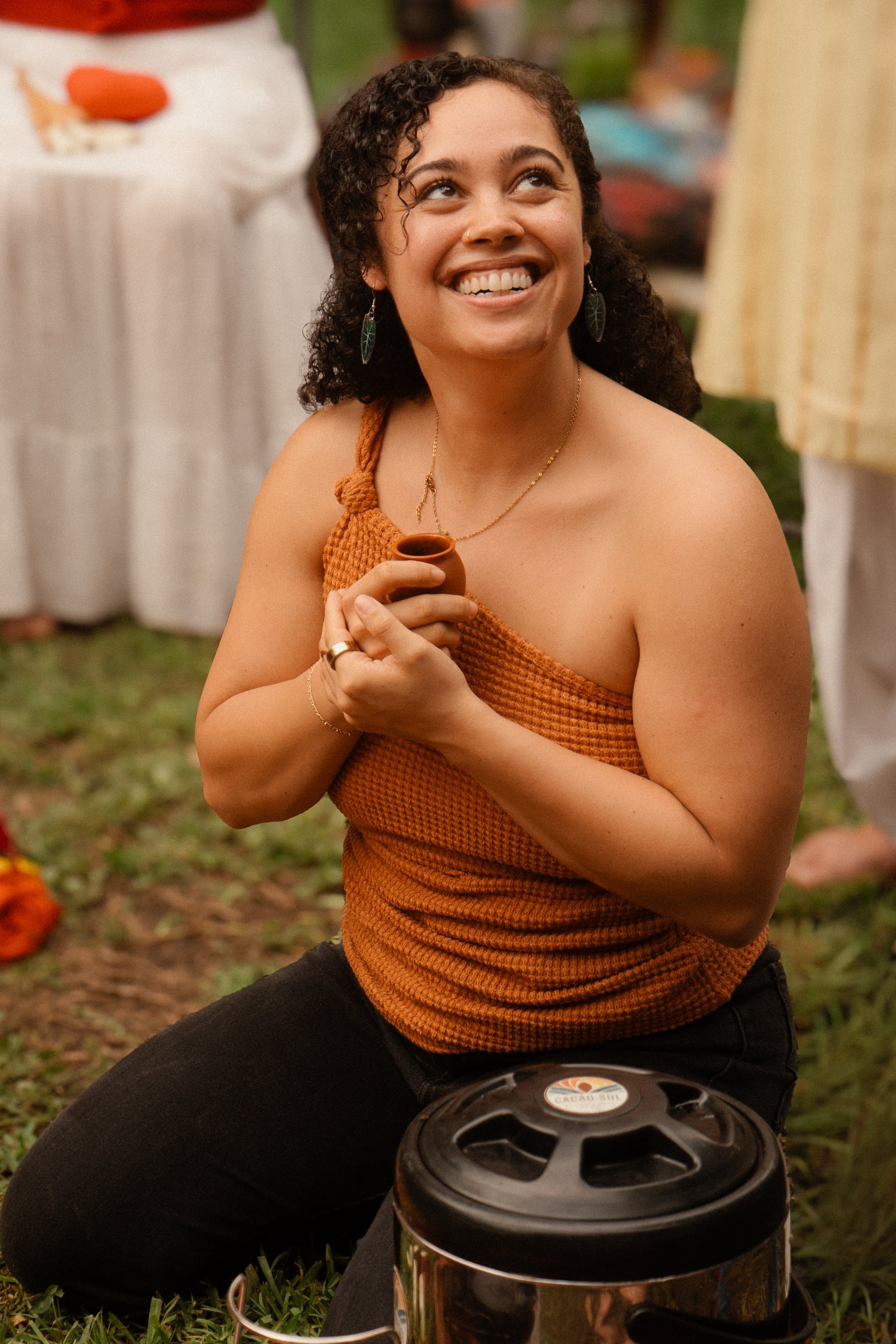 Woman in an orange top sitting on the grass with a metal container in front of her.